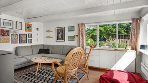Een gezellige woonkamer met een grijze bank, een klein rond tafeltje en rieten stoelen. Er hangen afbeeldingen in lijsten aan de muren, een raam met gordijnen en een uitzicht op groen.
