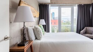 Une chambre d’hôtel avec un grand lit, deux lampes de chevet, une tête de lit en bois, une petite table avec une lampe et une vue sur la ville grâce à de grandes fenêtres.