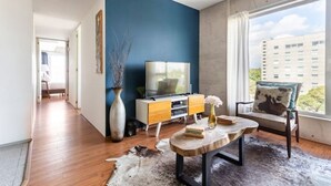 Un salon moderne avec un mur d’accent bleu, un téléviseur, une table basse en bois et une vue sur la ville.