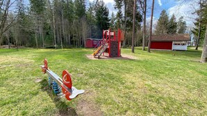Leikkikentällä on liukumäki, kiipeilyseinä ja keinut. Se sijaitsee nurmialueella, jossa on puita, ja taustalla näkyy punainen rakennus.