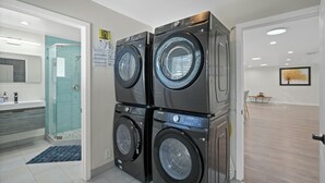 Une buanderie équipée d’une machine à laver et d’un sèche-linge à chargement frontal superposés, d’une cabine de douche en verre et, au fond, d’une petite table avec des chaises.