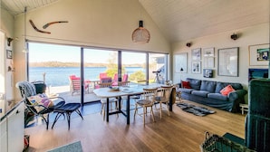 Un salón amplio con una mesa grande y sillas, un sofá y vistas a un lago a través de amplias ventanas.