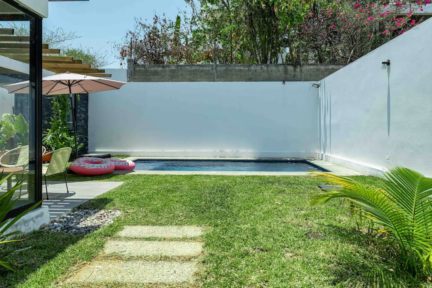 A small backyard with a swimming pool, a glass door leading to a patio area, and a concrete wall.