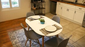 A dining area with a white table set for two, surrounded by chairs, a small potted plant, and a kitchen in the background. - Stone Cottage (Laurel Park)