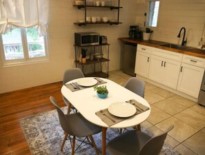 A dining area with a white table set for two, surrounded by chairs, a small potted plant, and a kitchen in the background.