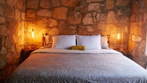 A bedroom with a stone wall, a bed with white bedding, and two wall-mounted lamps. - Stone Cottage (Laurel Park)