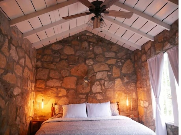 A bedroom with a stone wall, a bed with white bedding, a ceiling fan, and two wall-mounted lamps.