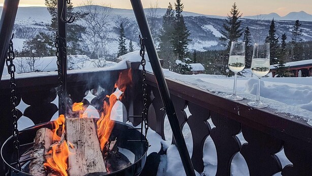 A fire pit with logs burning, surrounded by a wooden railing. In the background, there are snow-covered trees and mountains.