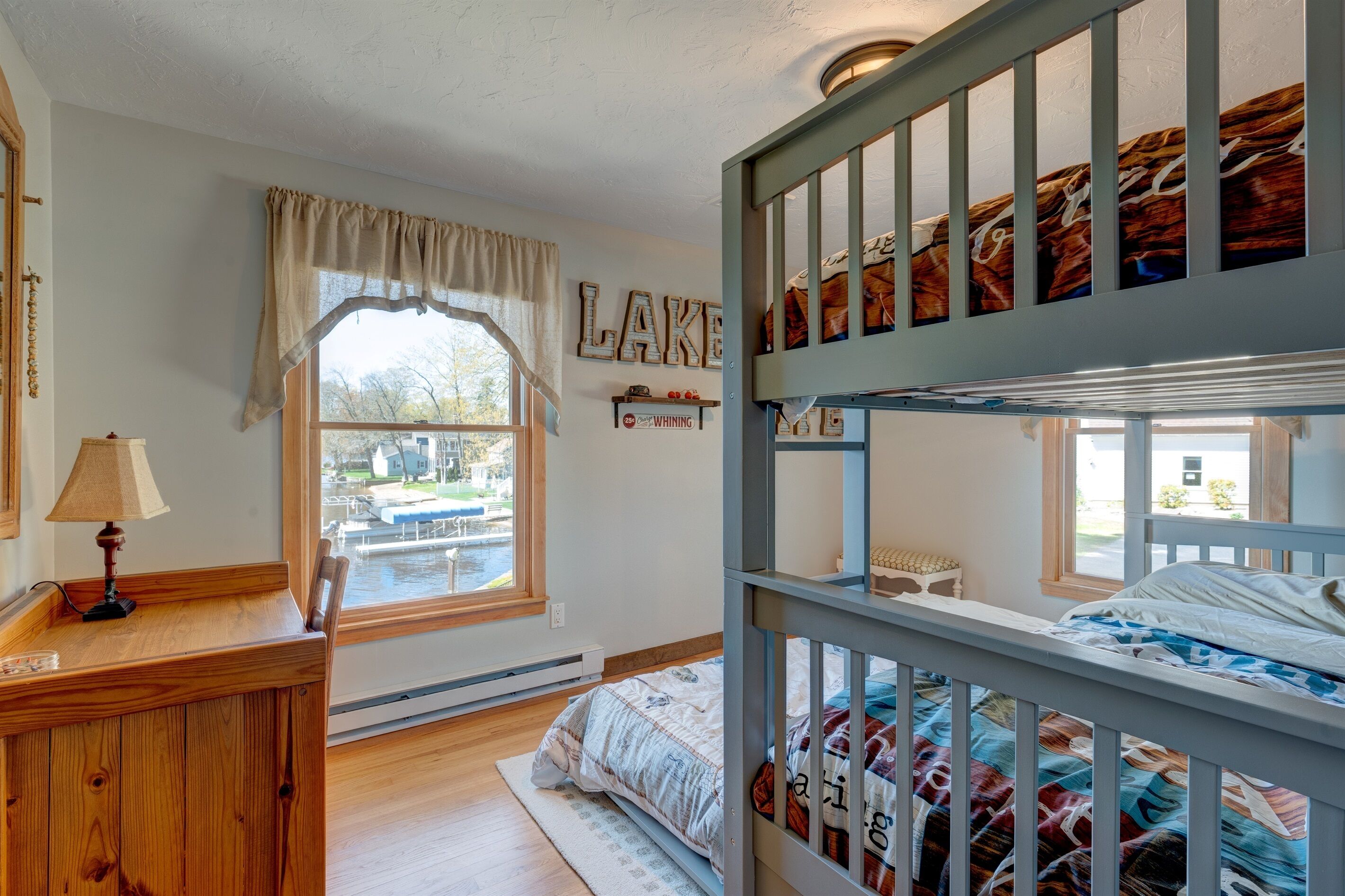 Habitación con literas y vistas a un puerto deportivo, un escritorio de madera con lámpara, y una estantería en la pared con adornos.