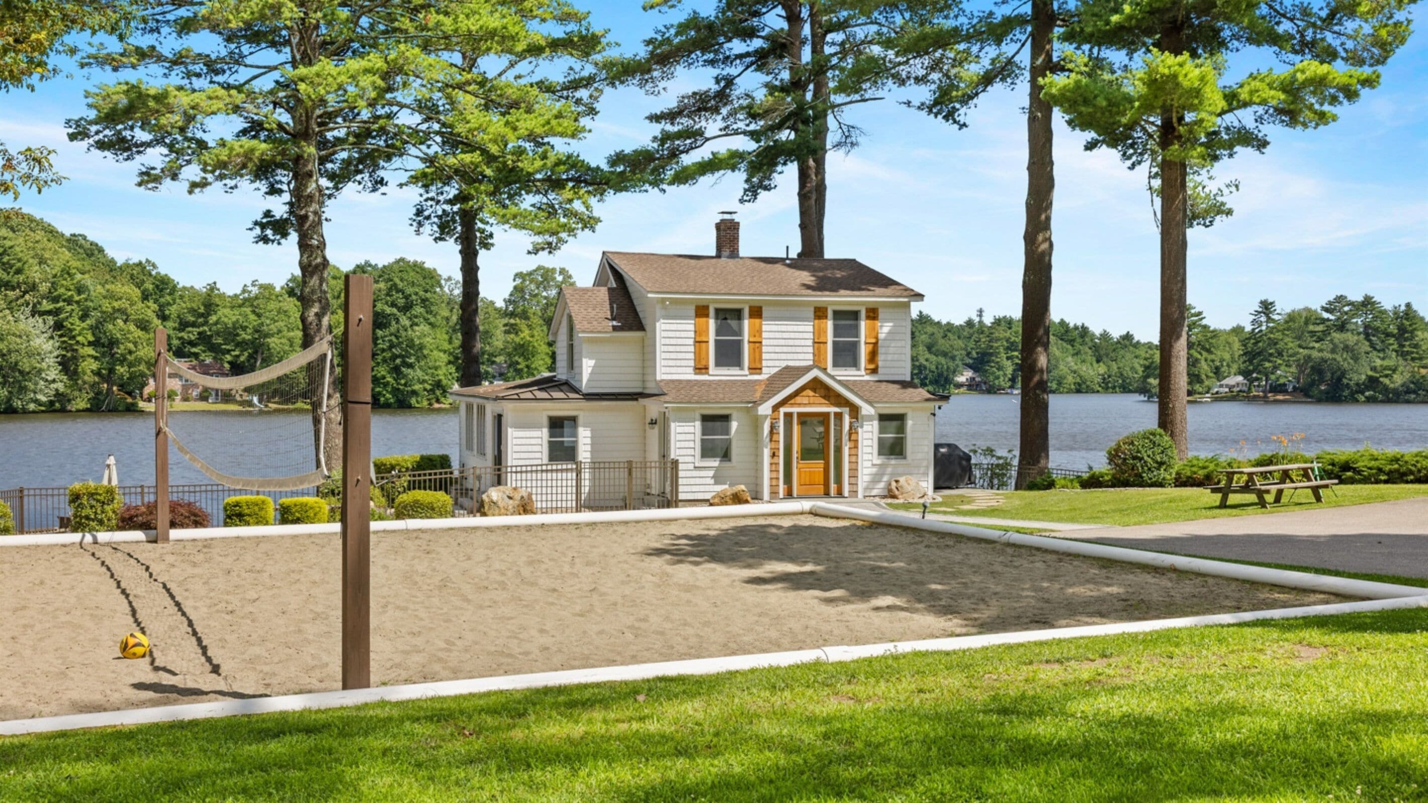 A two-story house with a sandy beach area in front, surrounded by trees and a lake.