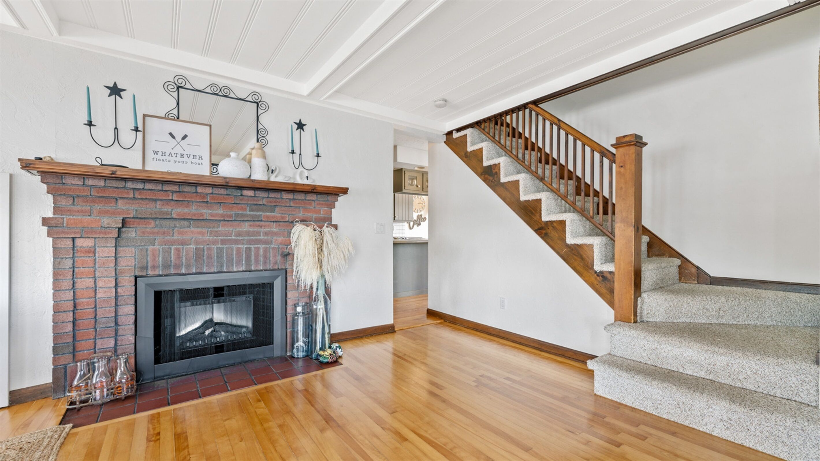 A spacious living room with a brick fireplace, wooden flooring, and a wooden staircase leading to an upper level.