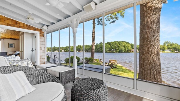 A spacious deck with wicker furniture, large windows offering a view of a lake and trees, and a wooden ceiling.