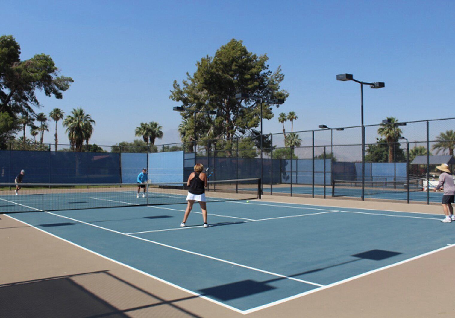 A tennis court with players, surrounded by a fence and palm trees.