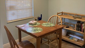 A dining area with a wooden table, two chairs, a window with blinds, a hanging light fixture, and a bookshelf with books and decorative items. - 🌴Tropical Cottage, In The Lovely Palm Beach Historic District LOCATION!  (West Palm Beach)