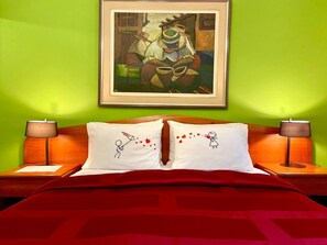 Una camera d'albergo con parete verde, un quadro incorniciato, due comodini con lampade, una coperta letto rossa e due cuscini con motivo a cuore.