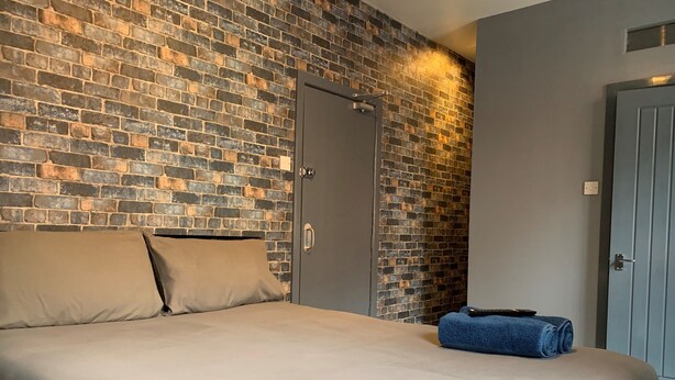 A bedroom with a brick-patterned wall, a bed with beige bedding, and a blue suitcase on a small table. - Urban Inn (Blackpool)