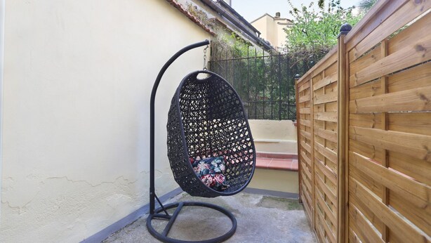 Een zwarte hangstoel met een bloemenkussen, een houten hek en een beige muur.