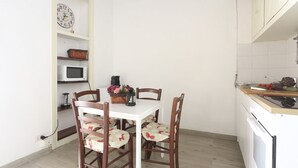 Een keuken met een witte tafel, vier stoelen, een magnetron, een mand en een klok aan de muur.