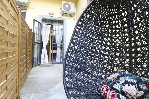 Un étroit couloir extérieur, délimité par une clôture en bois, avec une chaise en osier noir agrémentée d’un coussin à motifs floraux, et une porte en verre avec des rideaux.