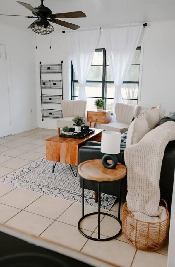 Un salon moderne avec un canapé, une table basse et un ventilateur de plafond. On y trouve une fenêtre avec des rideaux, une petite table d’appoint et un panier.