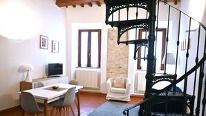 A dining area with a table and chairs, a television, a white armchair, and a black spiral staircase. - S318 - Sirolo, wonderful three-room apartment in a historic building (Sirolo)