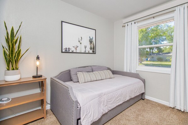 A bedroom with a grey sofa bed, a bedside table with a lamp, a framed picture on the wall, and a window with curtains. - Tourists’ Haven~Clean~Centrally Located (Rapid City)