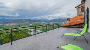 Un balcon donnant sur une ville et des montagnes, avec des chaises vertes et un bâtiment en pierre surmonté d’un toit rouge.