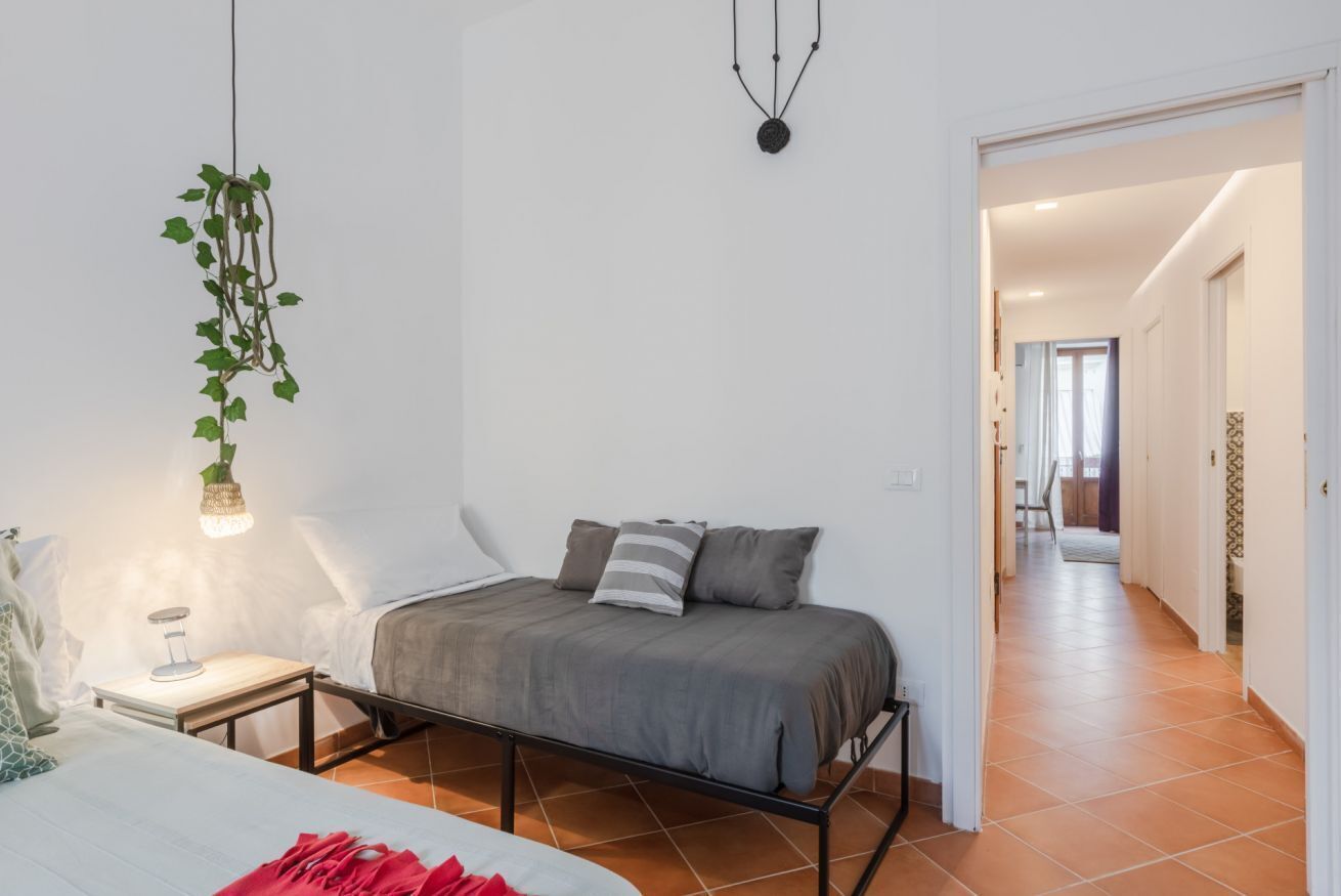 A bedroom with a bed, bedside table, lamp, hanging plant, and a view into the hallway.