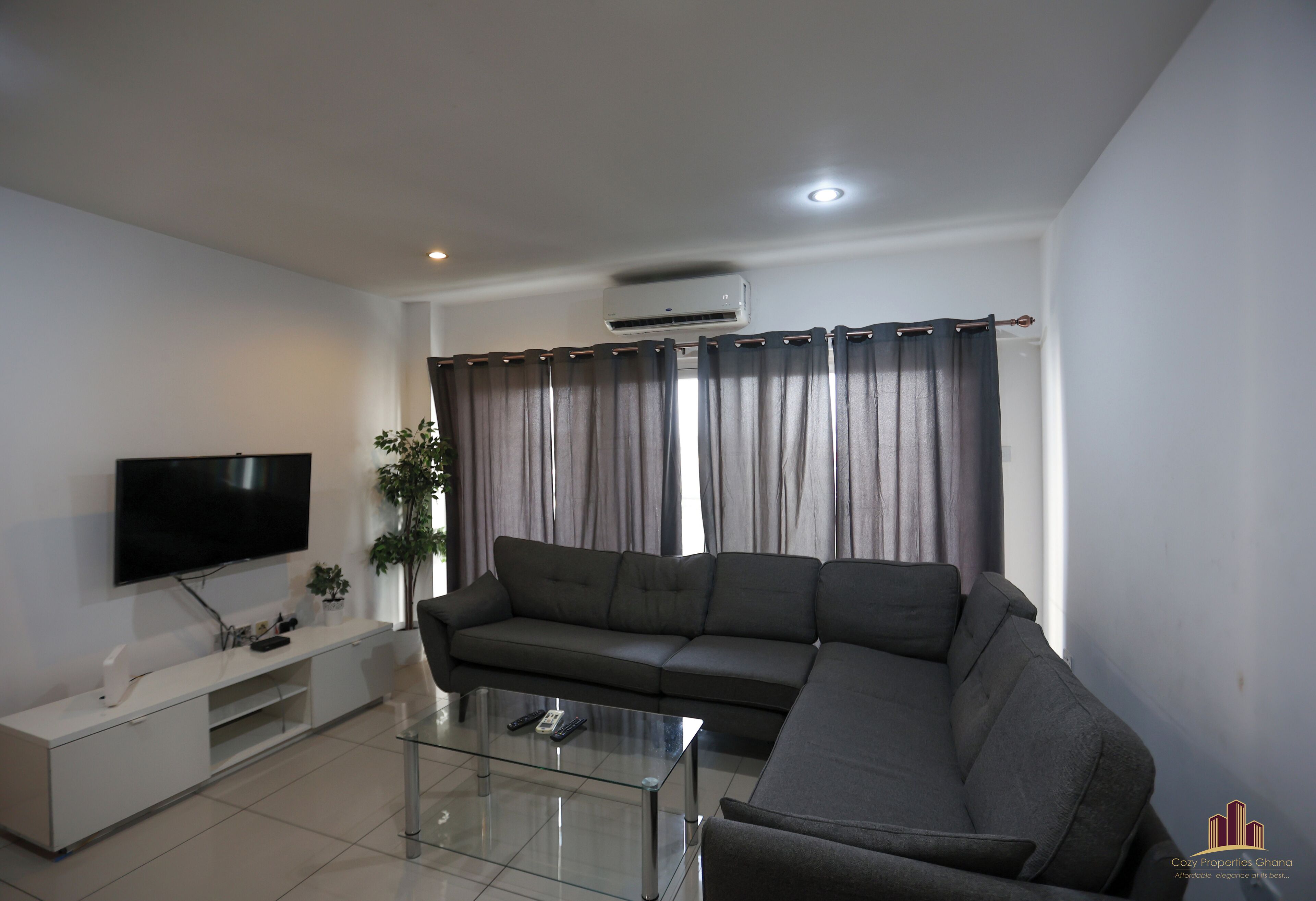 A modern living room with a flat-screen TV, a grey sofa, a glass coffee table, and a white entertainment unit.