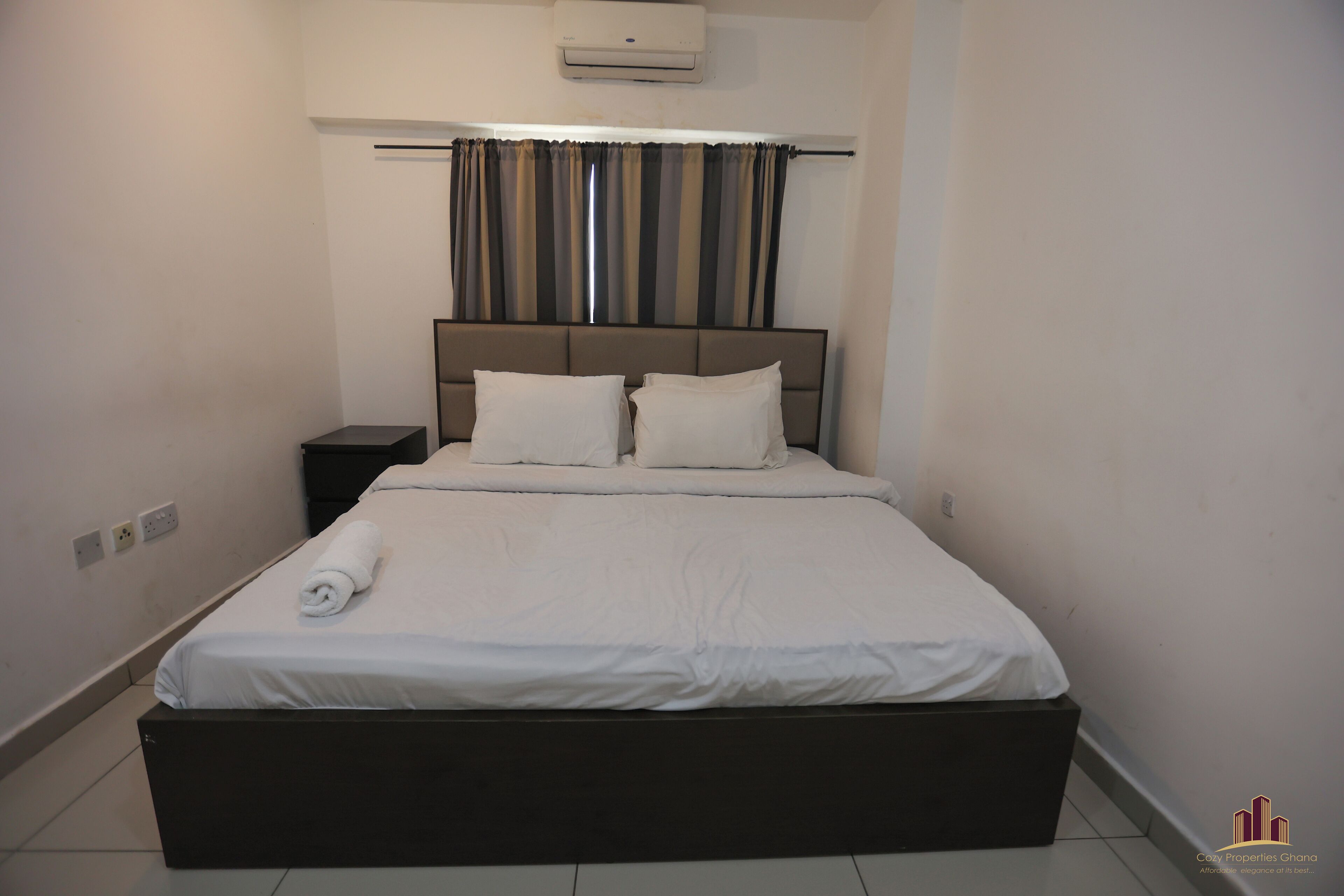 A neatly made bed with a dark headboard, a small bedside table, and a wall-mounted air conditioner.