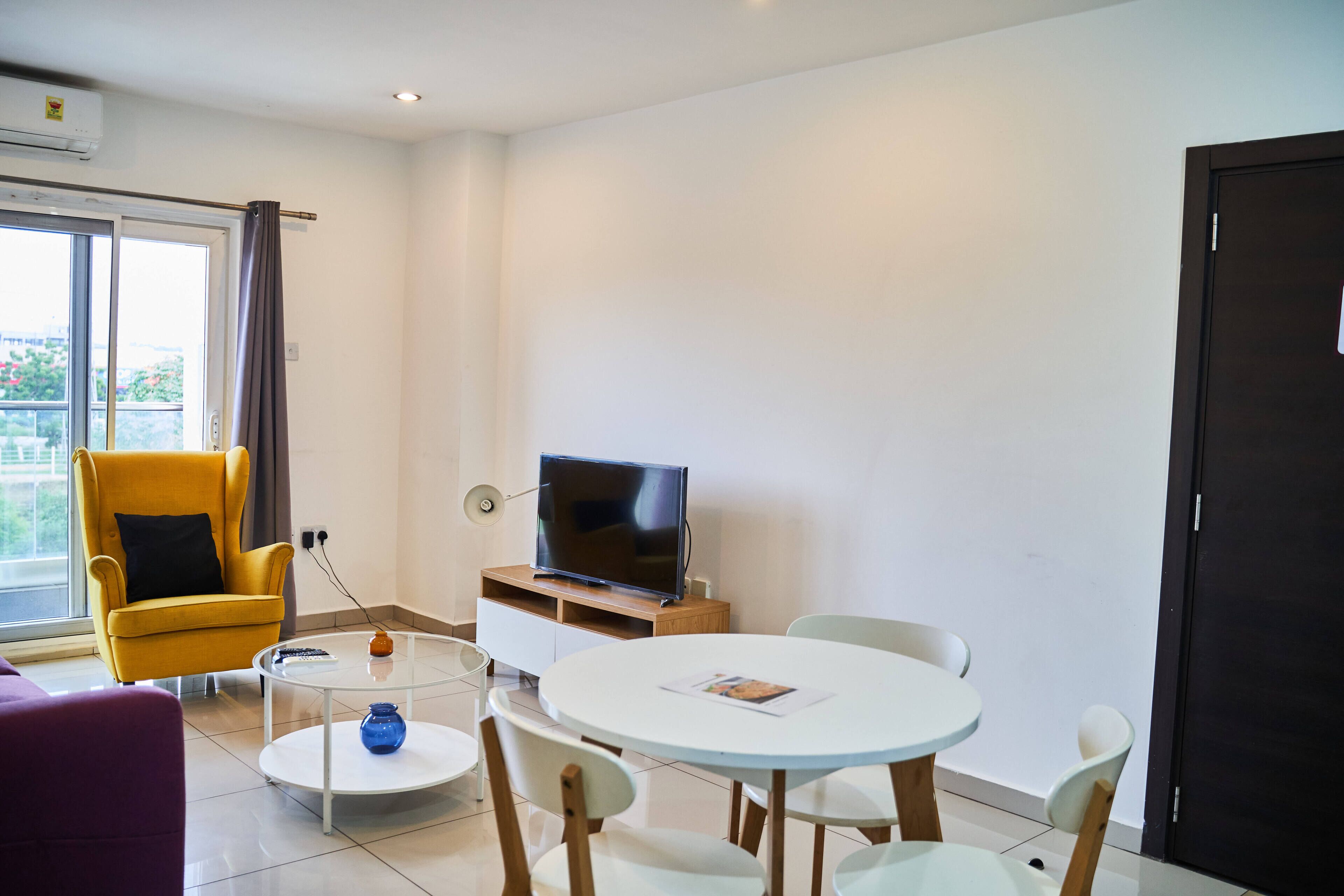 Een moderne woonkamer met een bank, een gele fauteuil, een ronde eettafel met stoelen, een flatscreen televisie op een houten dressoir en een raam met gordijnen.