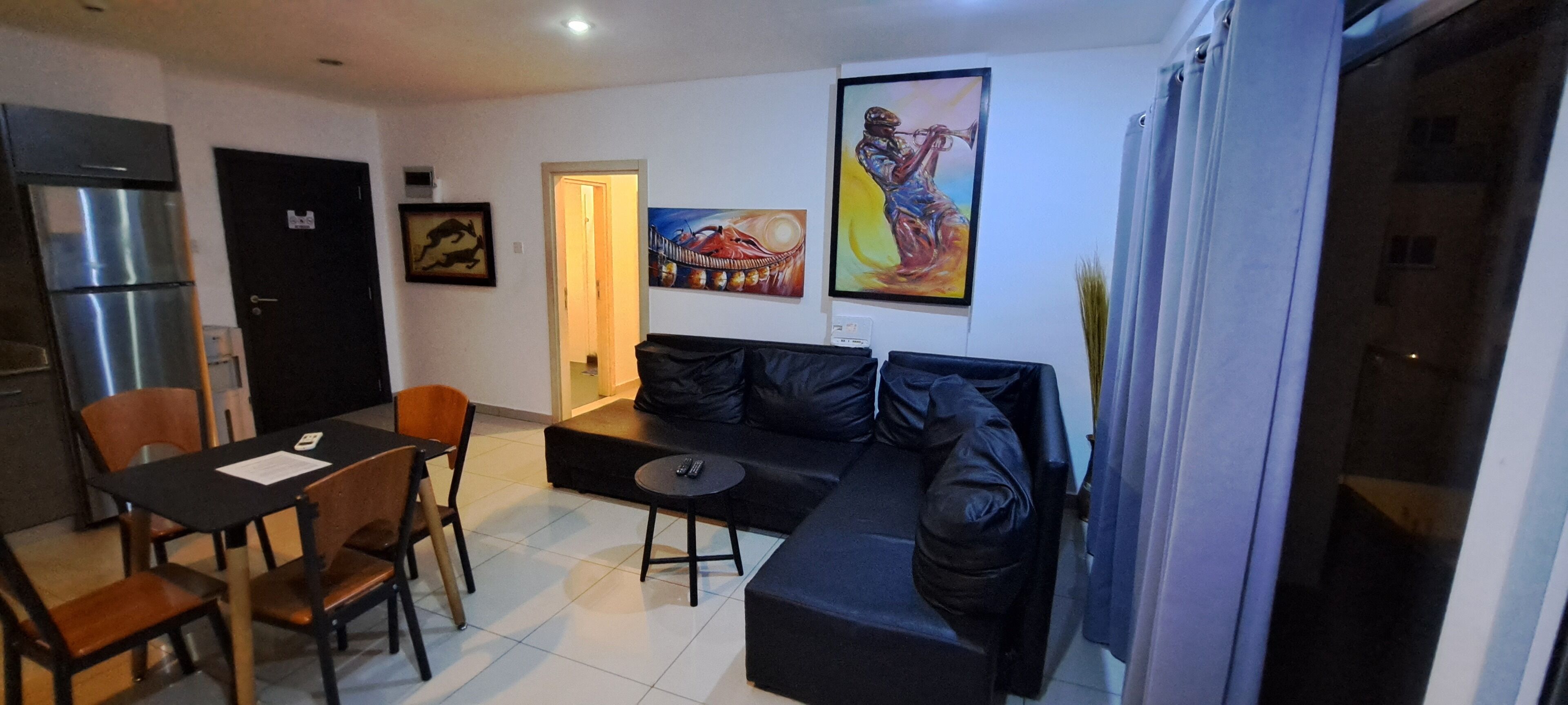 Un salon avec un canapé noir, une table à manger avec des chaises, un réfrigérateur et un tableau accroché au mur.