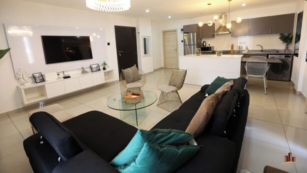 A modern living room with a black sofa, glass coffee table, and a TV mounted on the wall. - Gallery by Cozy (Accra)