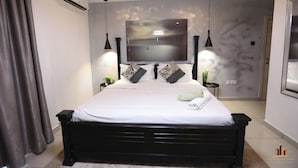 A modern bedroom with a large bed, a flat-screen TV mounted on the wall, pendant lights, and a small side table with a plant. - Gallery by Cozy (Accra)