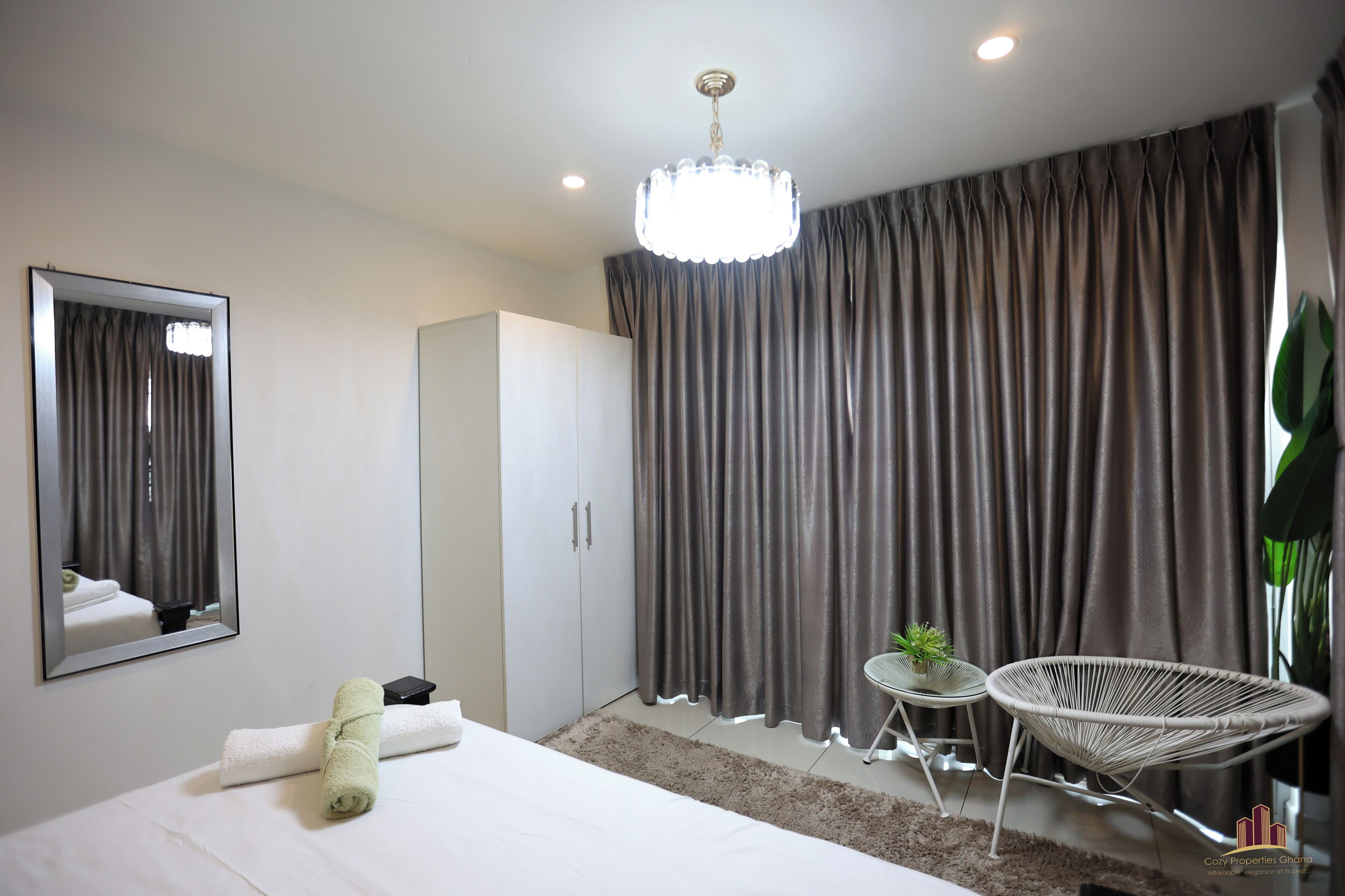 Een moderne hotelkamer met een groot bed, een spiegel, een kledingkast, een klein tafeltje met een plant en een rieten stoel.
