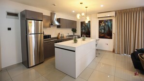A modern kitchen with a stainless steel refrigerator, a central island, and a dining area with a black sofa. - Gallery by Cozy (Accra)