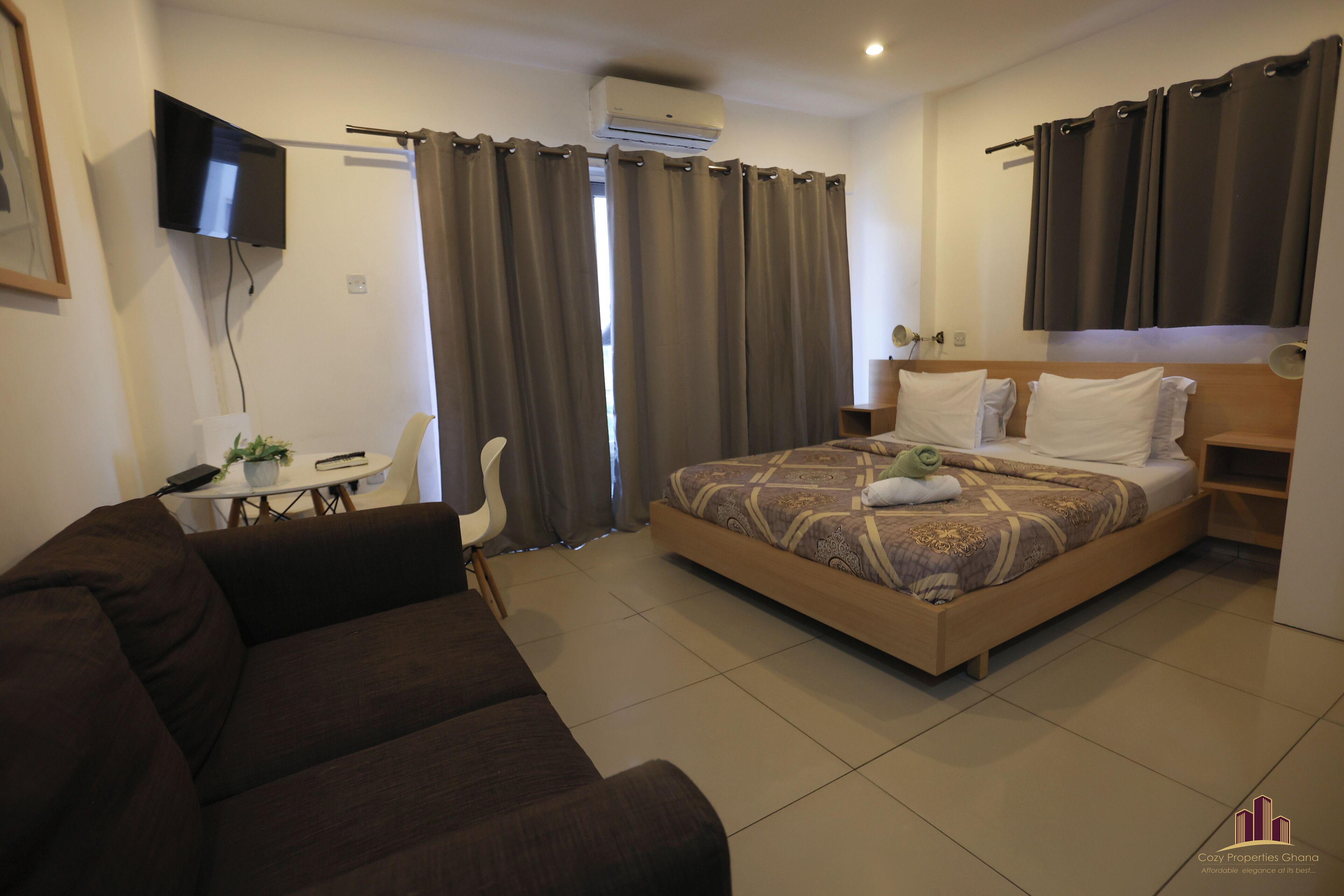 A hotel room with a bed, a TV mounted on the wall, a desk with a chair, and a couch.