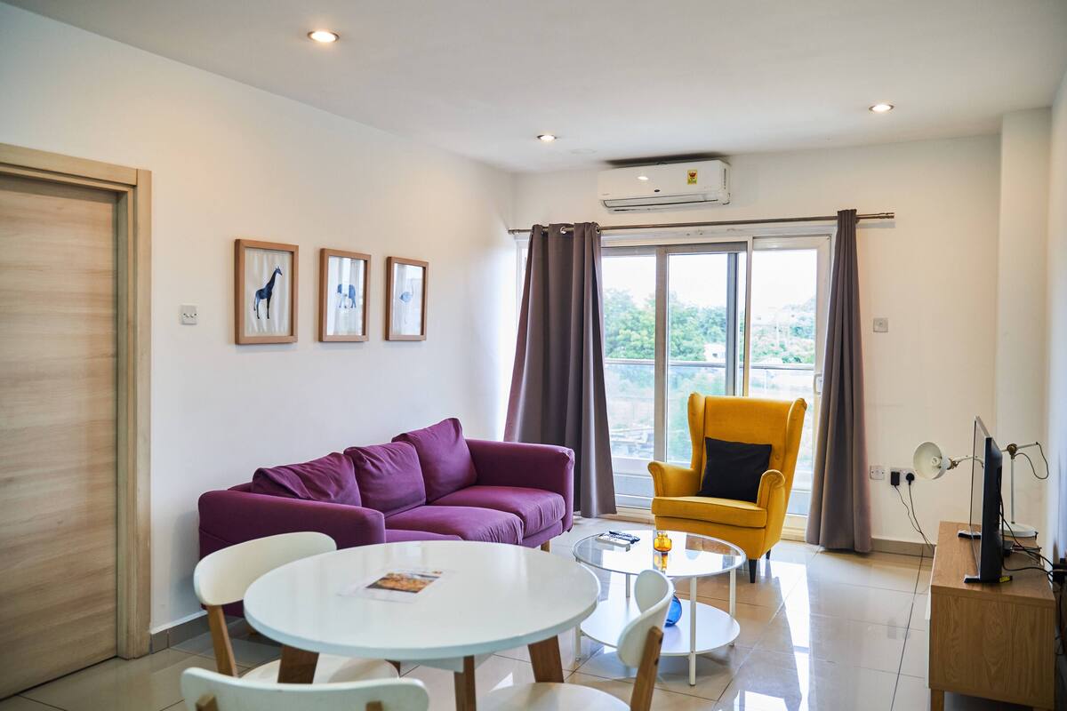 A modern living room with a purple sofa, a yellow armchair, and a white table. There are three framed pictures on the wall, a window with curtains, and an air conditioning unit on the ceiling.