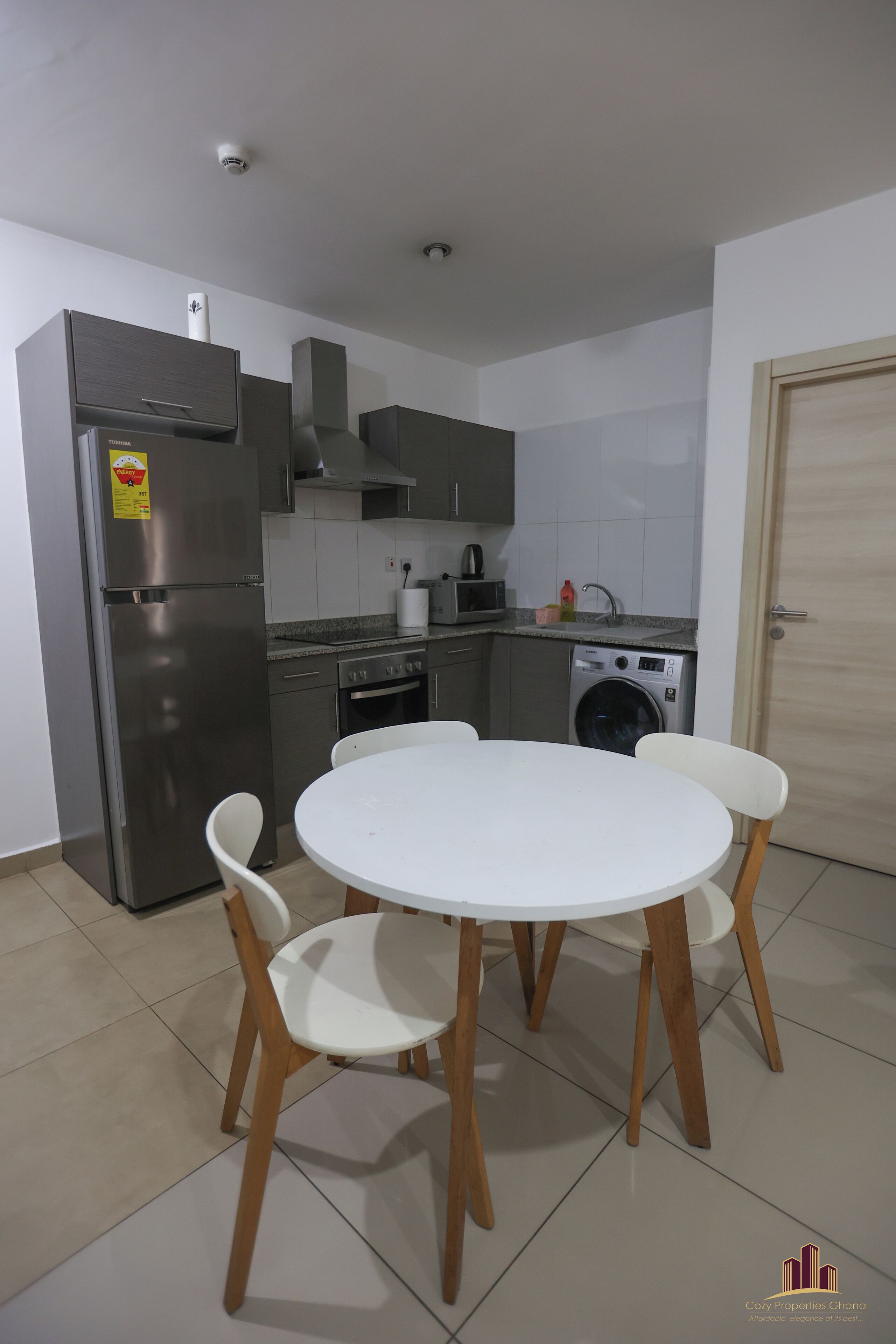 A compact kitchen with a refrigerator, microwave, oven, and washing machine. There is a round table with four chairs.