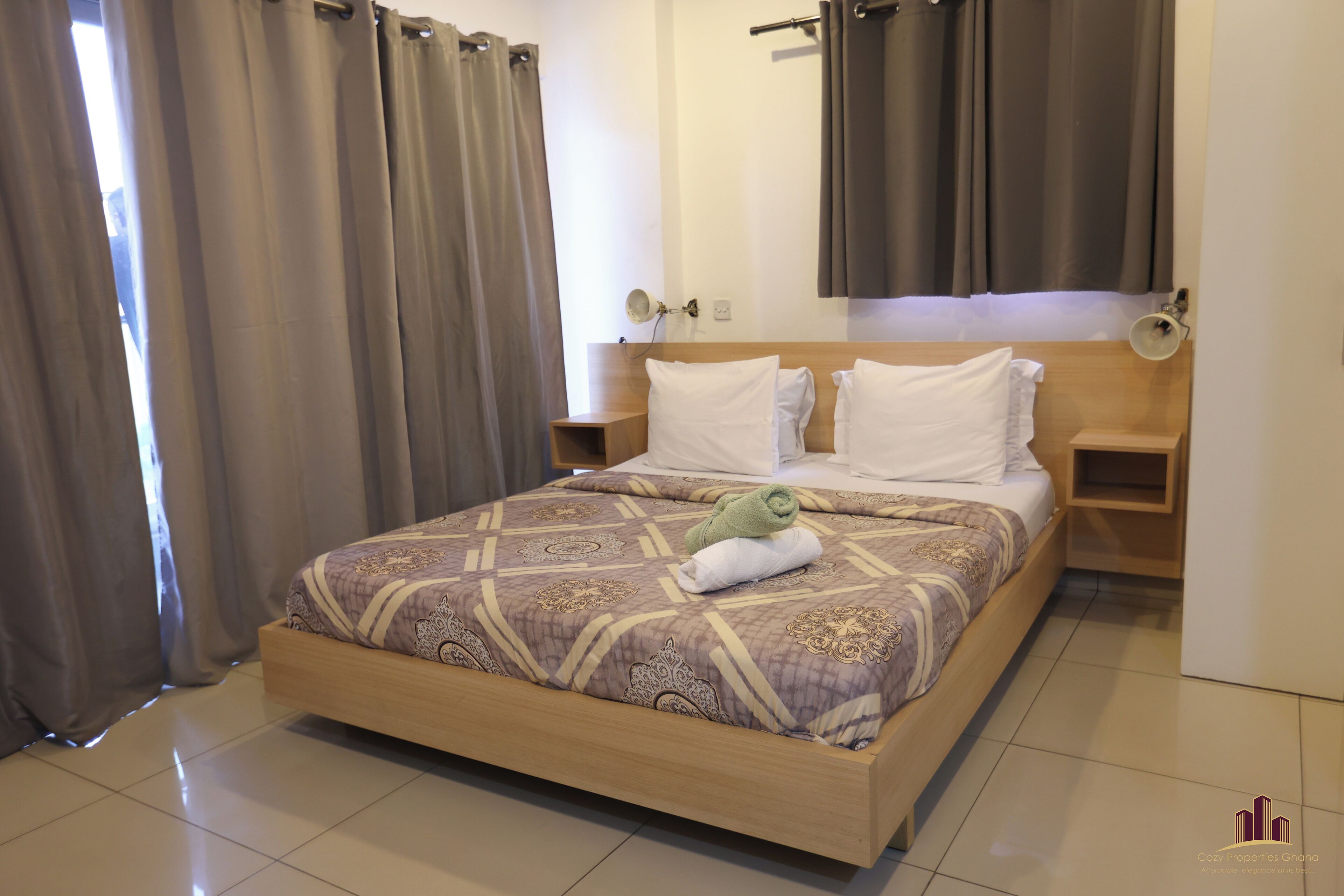 A neatly made bed with a patterned bedspread, two pillows, and a rolled towel. There are two bedside tables with lamps, and a wooden headboard.
