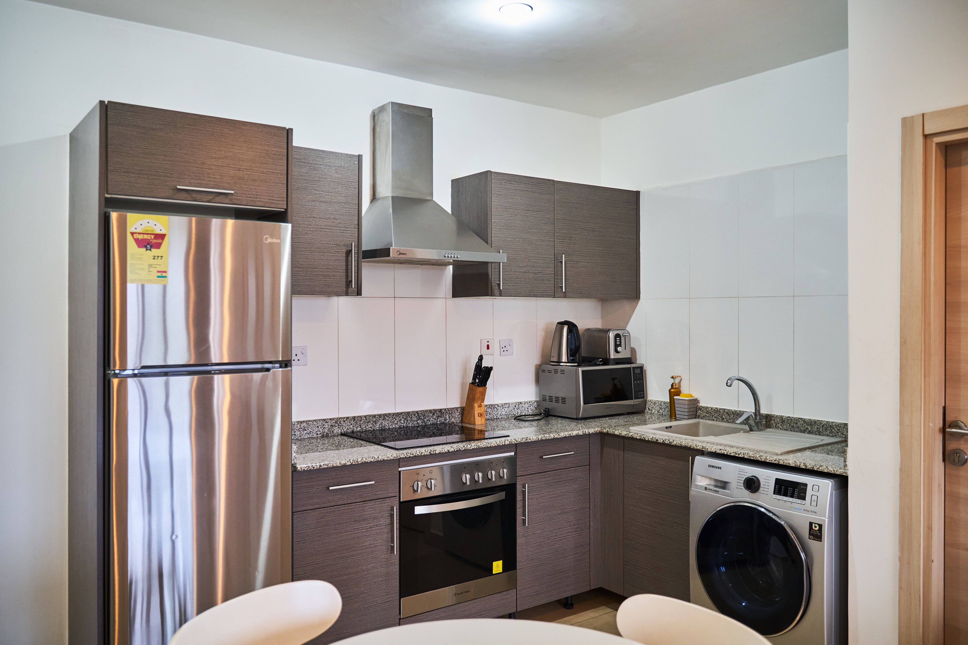 A compact kitchen with a stainless steel refrigerator, built-in oven, microwave, and washing machine.