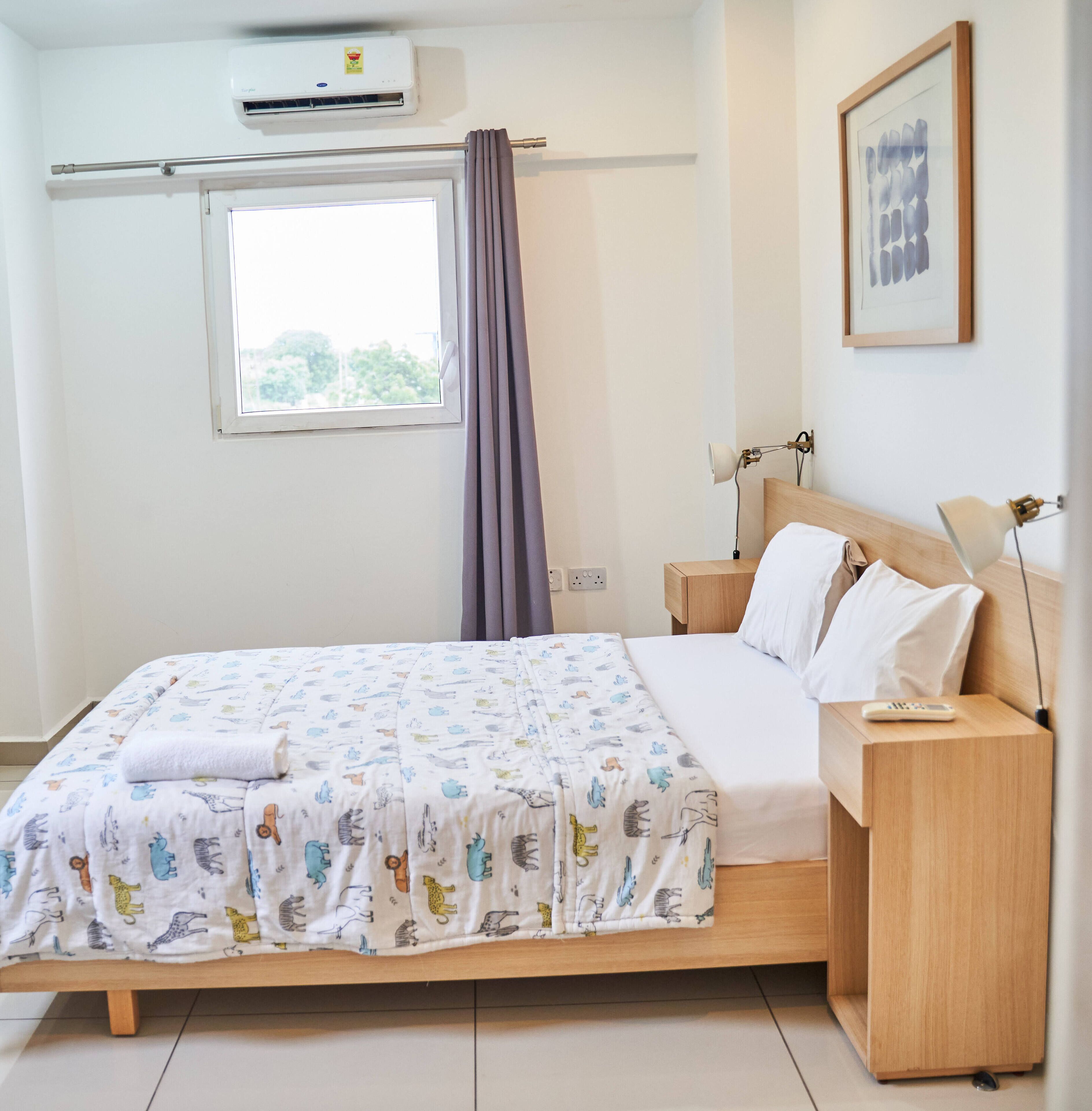 A bedroom with a bed, bedside tables, a lamp, an air conditioner, a window with curtains, and a framed picture on the wall.