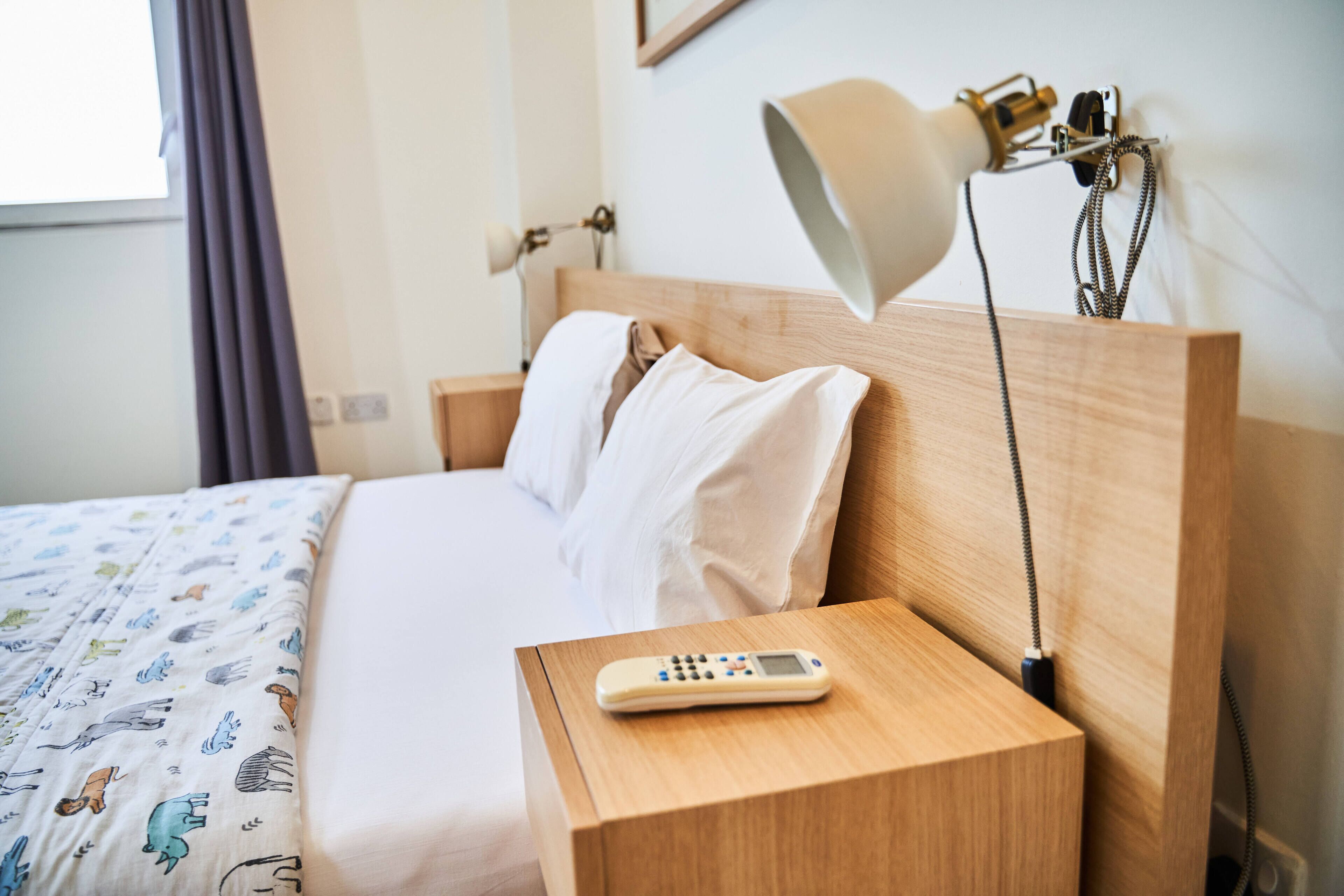 Una cama bien hecha con un edredón estampado, una lámpara de noche y un control remoto sobre una mesita de madera.