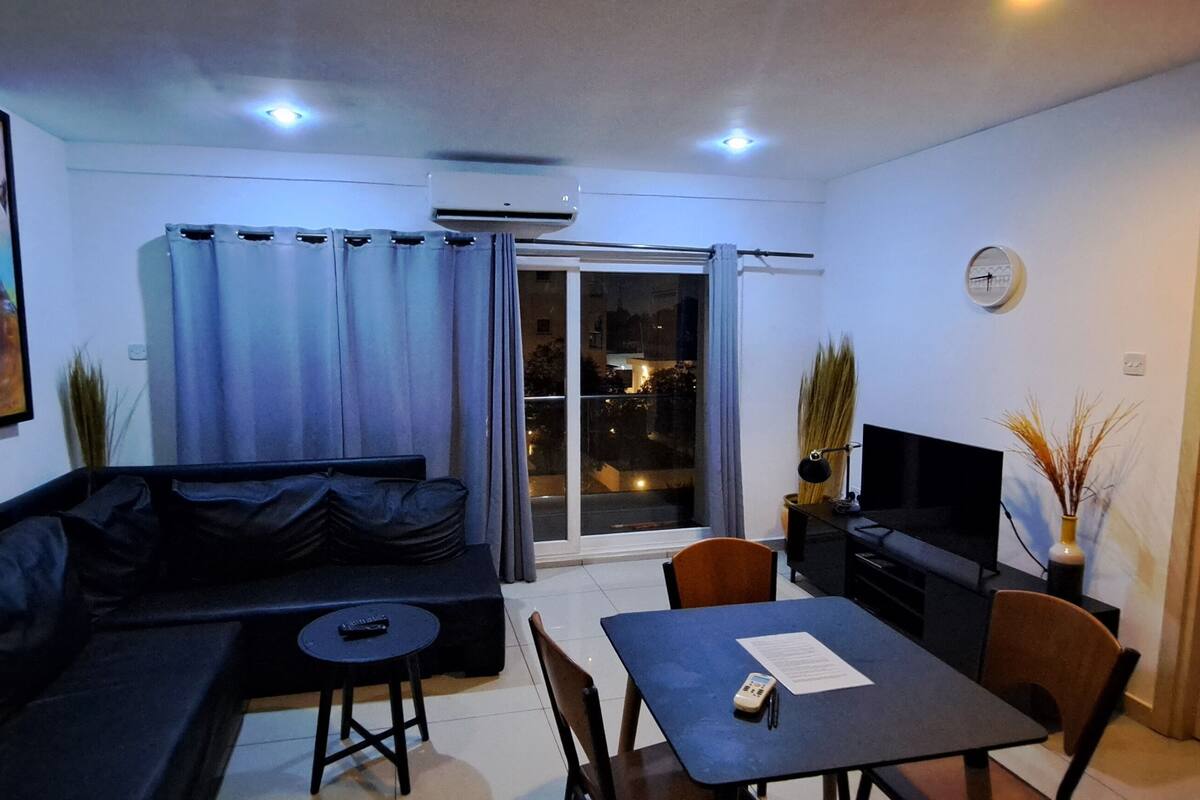 A compact living space with a sofa, a small dining table, a TV, and a sliding glass door leading to a balcony.