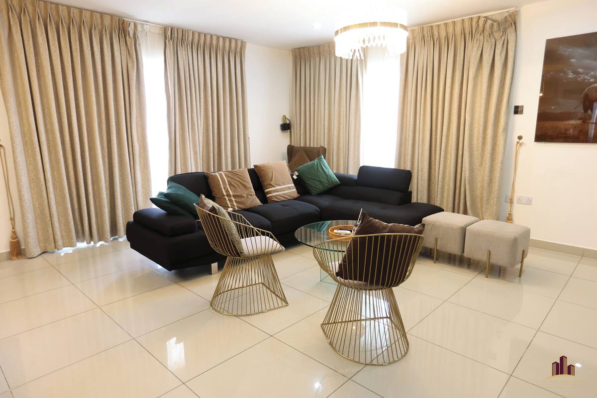 A modern living room with a black sectional sofa, two gold wire chairs, a glass coffee table, and beige curtains.