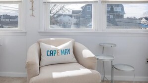 A cozy room with a beige armchair, a small round ottoman, and a white side table. A window with white blinds offers a view of houses outside. - 4 Blocks to Beach, Linens Provided (Cape May)