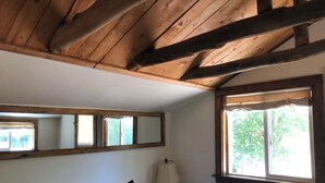 A bedroom with a wooden ceiling, a bed with a white and brown patterned bedspread, a bedside lamp, and a window with a view of greenery. - Newly Restored Vintage Cabin near Bryce, Grand Staircase. Great views! (Henrieville)