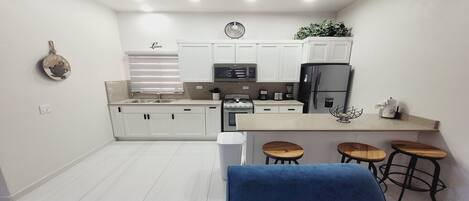 A modern kitchen with white cabinets, a stainless steel refrigerator, a microwave, and a gas stove. There is a bar area with stools and a blue towel.