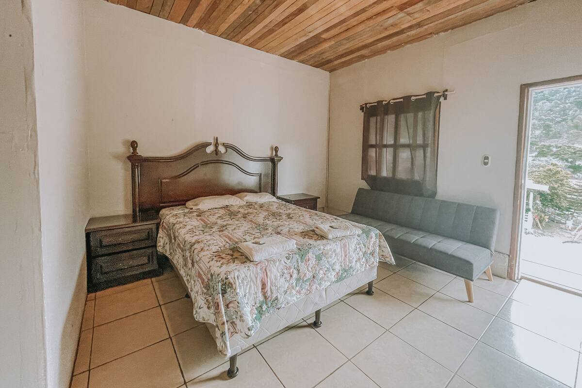 Ein Schlafzimmer mit Holzbalkendecke, einem Bett mit floralem Überwurf, einem Nachttisch, einer Bank und einem Fenster mit Vorhängen.