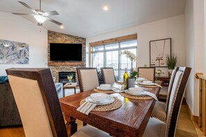 A dining area with a wooden table set for four, surrounded by chairs, a television mounted on a stone wall, and a large window with a view.